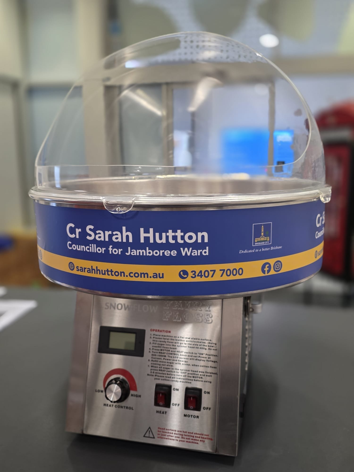 A cotton candy machine displaying a bumper sticker with the name and contact information of Cr Sarah Hutton, a councillor for Jamboree Ward. The sticker also includes a website, phone number, and social media icons.