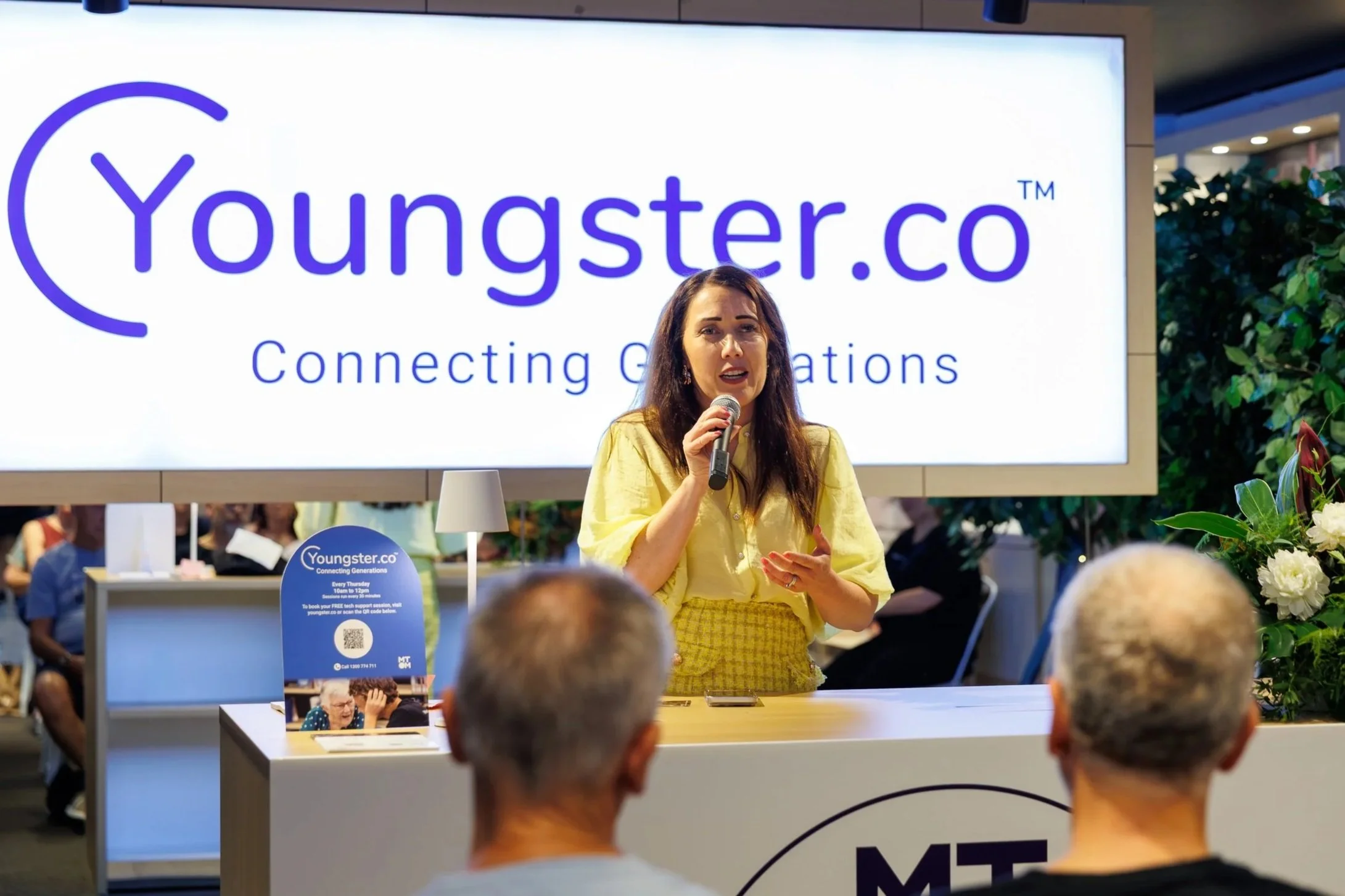 Cr Sarah Hutton holding a microphone speaking at a conference, with a large screen behind her displaying the logo and tagline of Youngster.co, 'Connecting Generations,' in a busy indoor setting with plants and attendees.