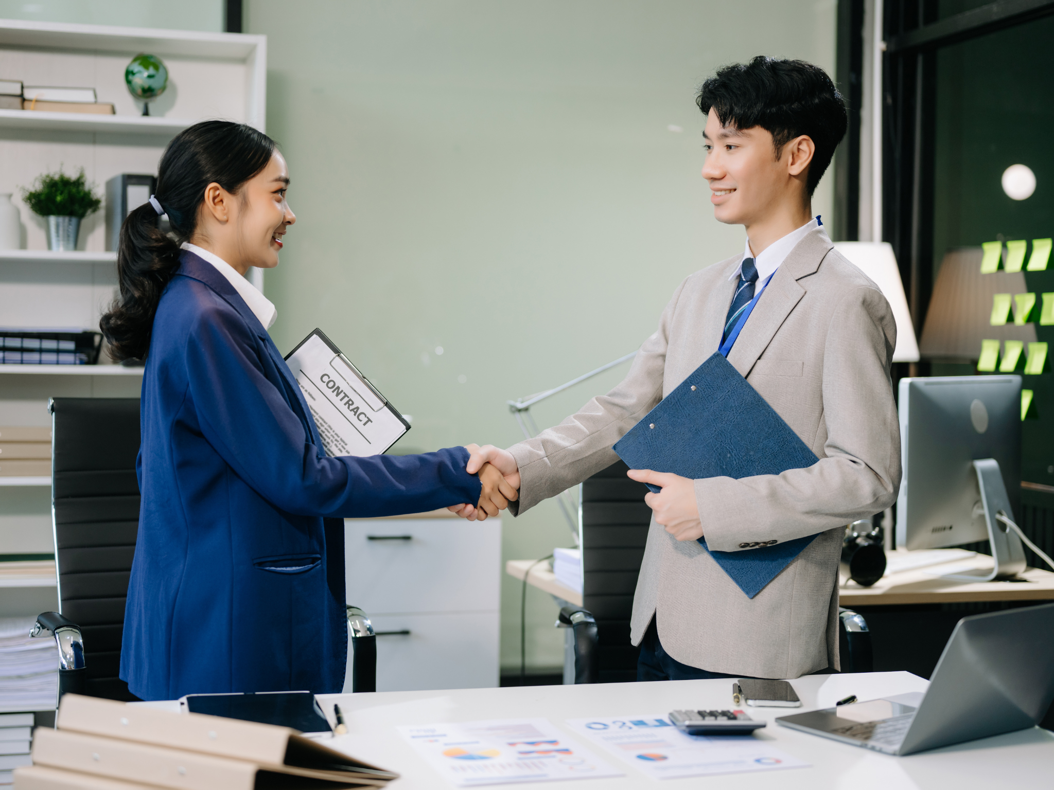 A man and woman shaking hands in an office, smiling, with documents and a computer on the desk.