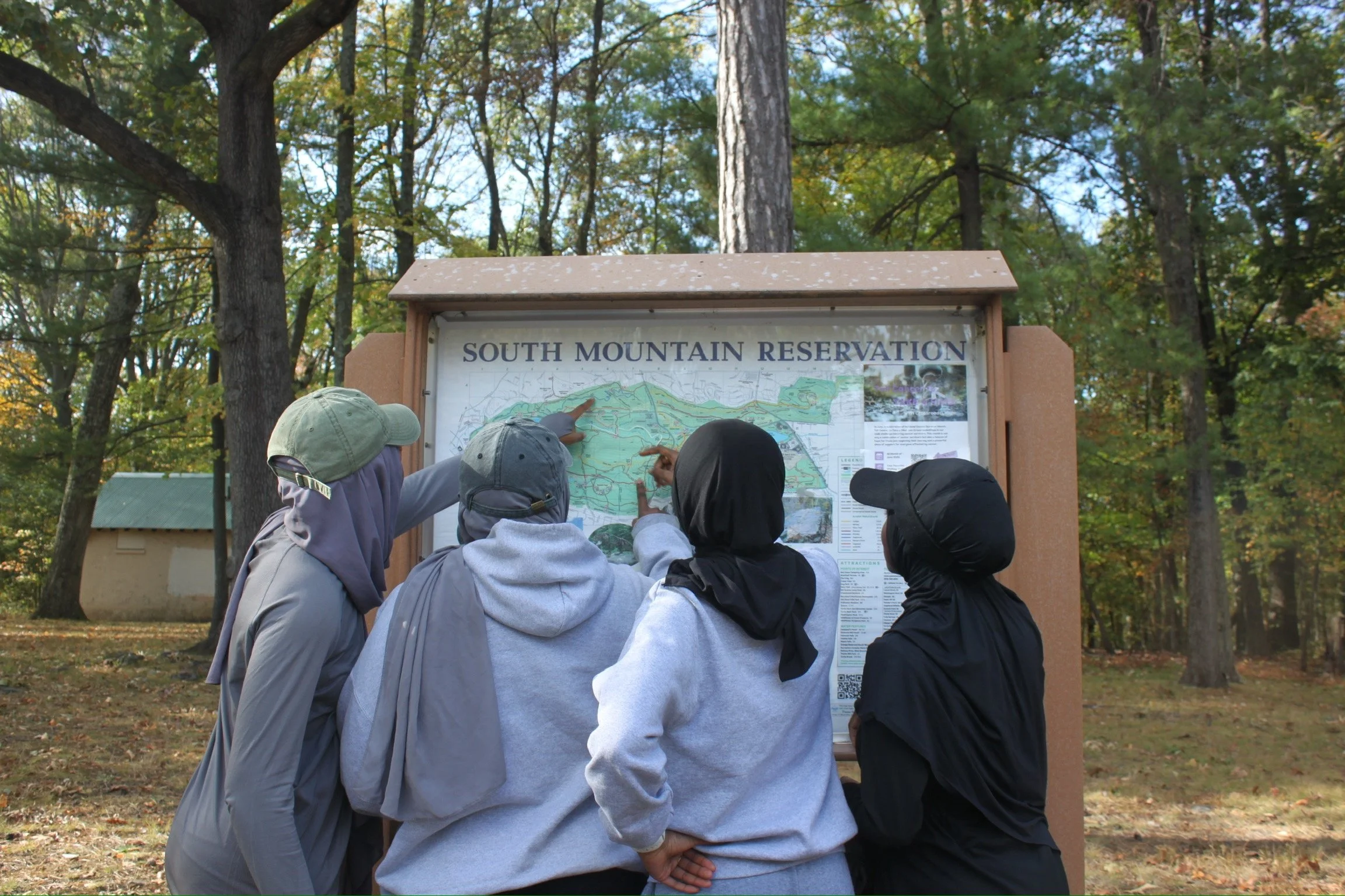 Four people gather around an informational map at South Mountain Reservation, with trees and a small building in the background.