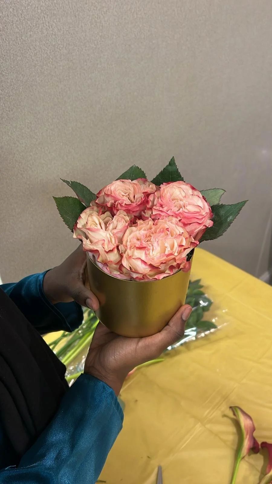 A person holding a gold-colored pot filled with pink and peach-colored roses with green leaves