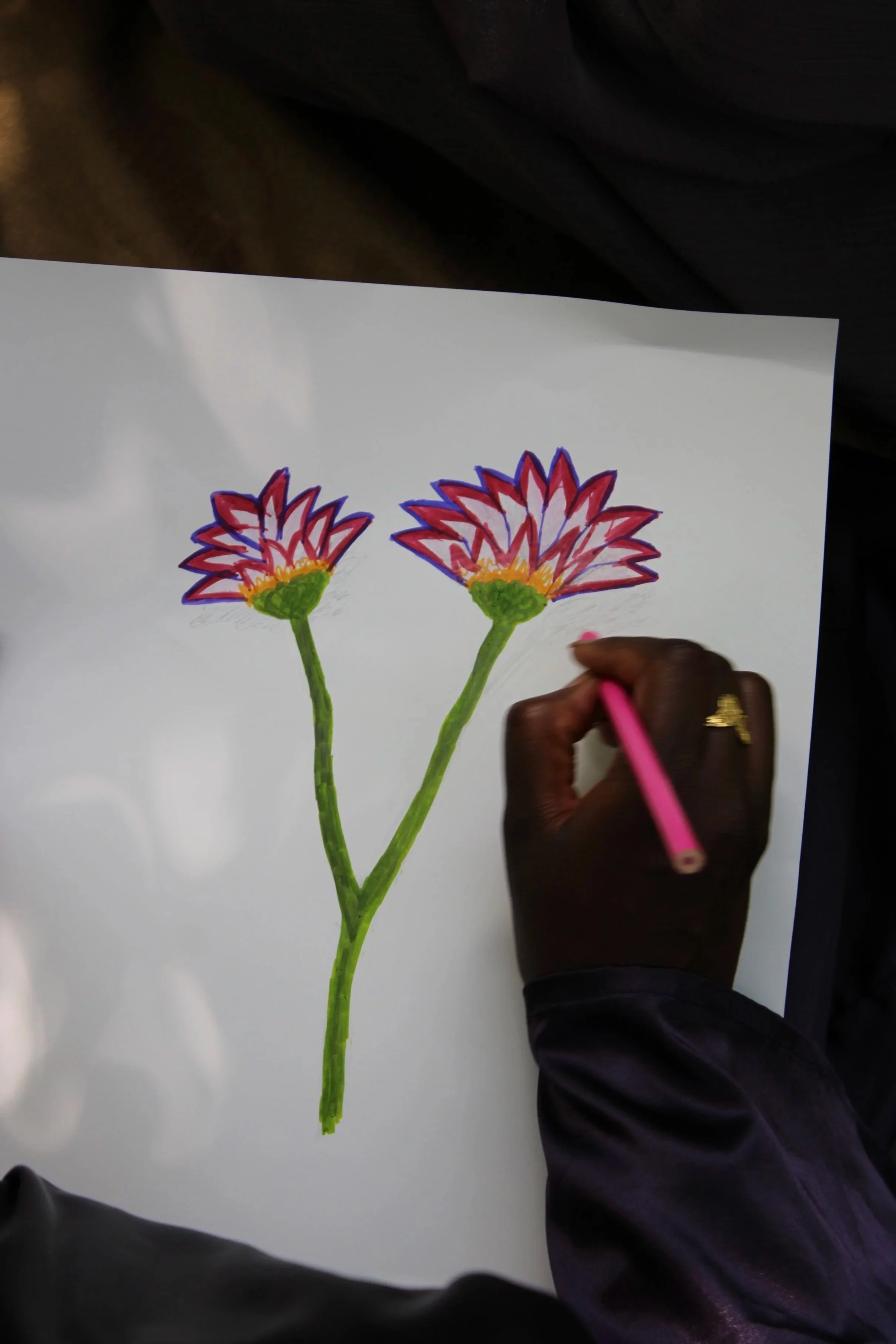 A person with dark skin is drawing two pink flowers with yellow centers and green stems on white paper using a pink colored pencil. The person is wearing a black long-sleeve garment and a gold ring.