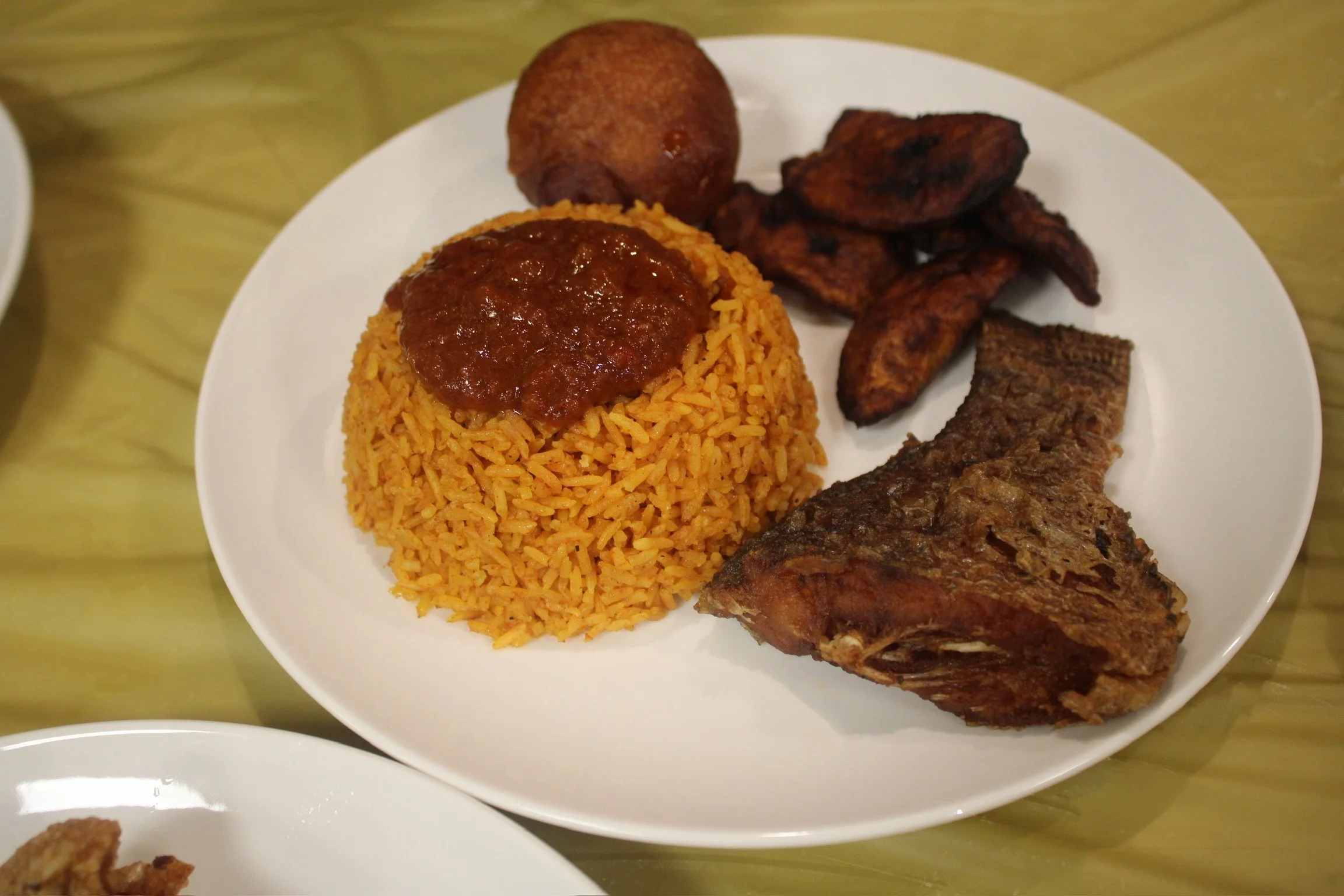 Plate of Nigerian dishes including rice with tomato sauce, fried plantains, a fried ball, and a piece of fried fish.