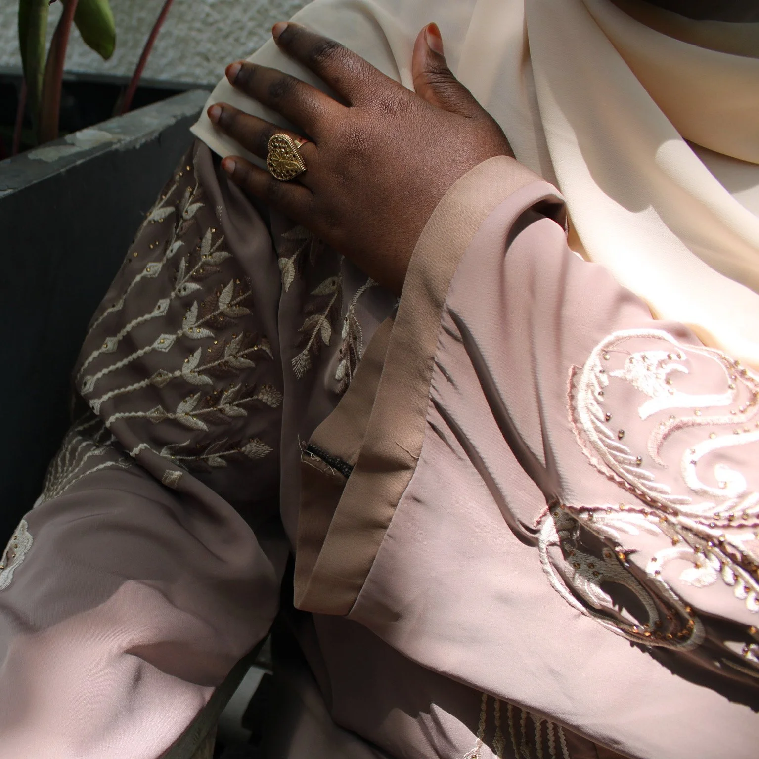 Close-up of a person wearing a beige traditional embroidered garment, showing their hand with a gold ring resting on their chest.