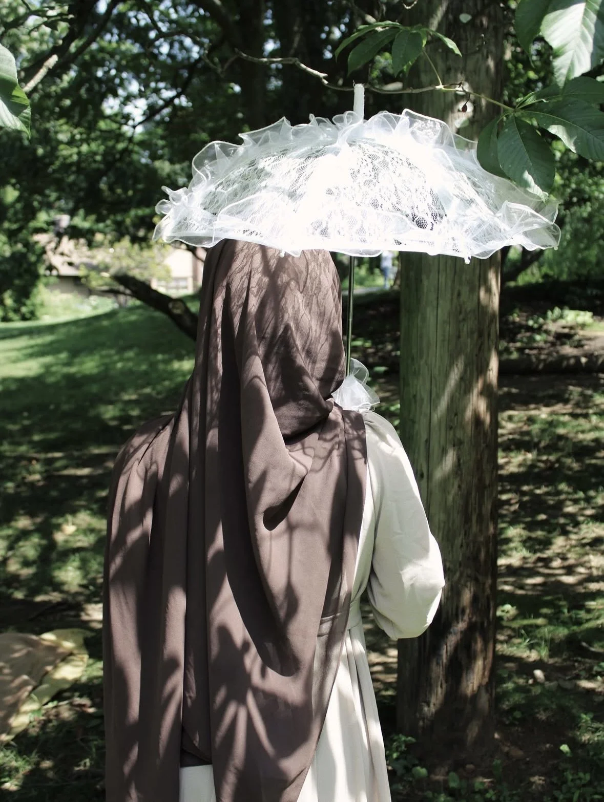 A person wearing a dark brown headscarf, a light beige coat, and a white dress, holding a white lace parasol with ruffled edges, standing in a shaded outdoor area with trees and grass.
