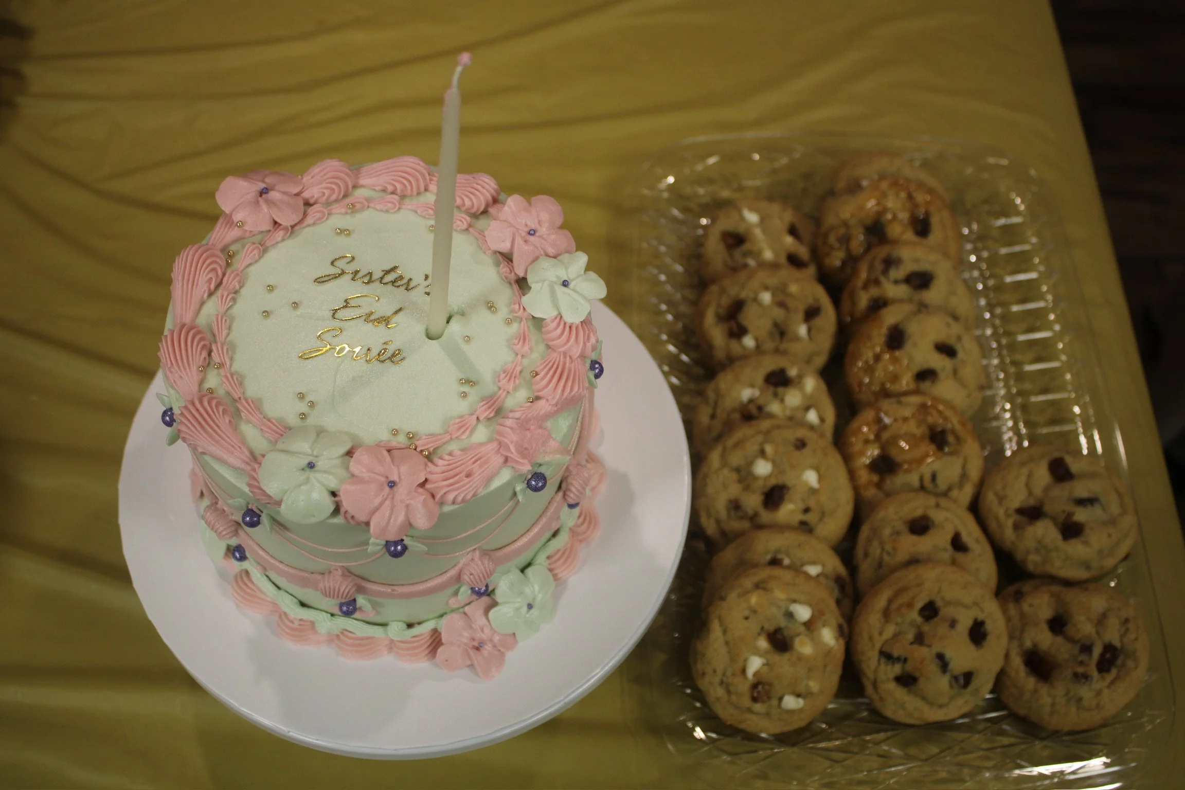 Decorated birthday cake with pink and white icing, flowers, and gold lettering, next to a tray of chocolate chip cookies on a yellow table.
