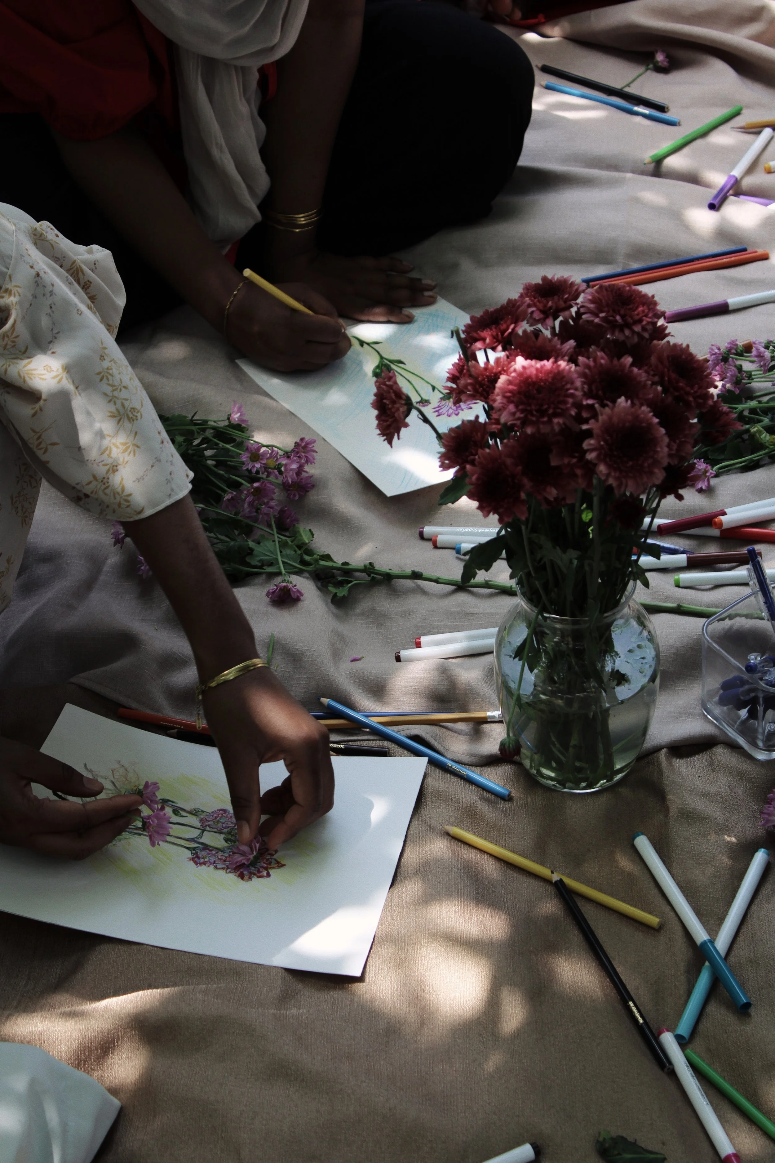 Hands of two people arranging pink and purple flowers on paper, surrounded by scattered colorful markers on a beige cloth.