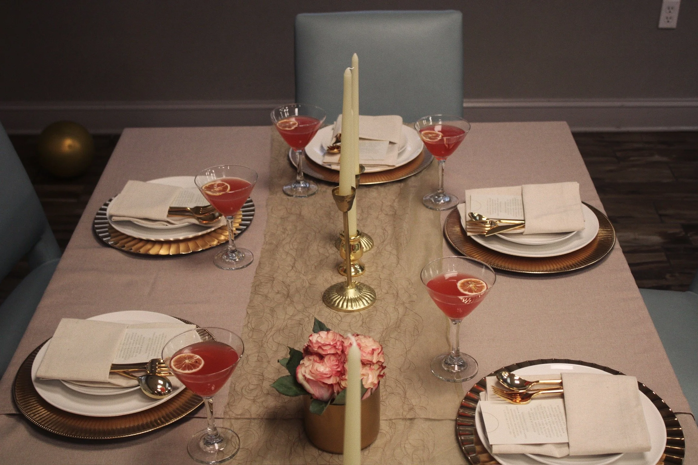 A formal dining table set for six with gold and silver accents, a centerpiece with pink flowers, a gold candle holder with a tall white candle, and glasses of pink cocktails, with napkins, plates, and cutlery arranged at each seat.