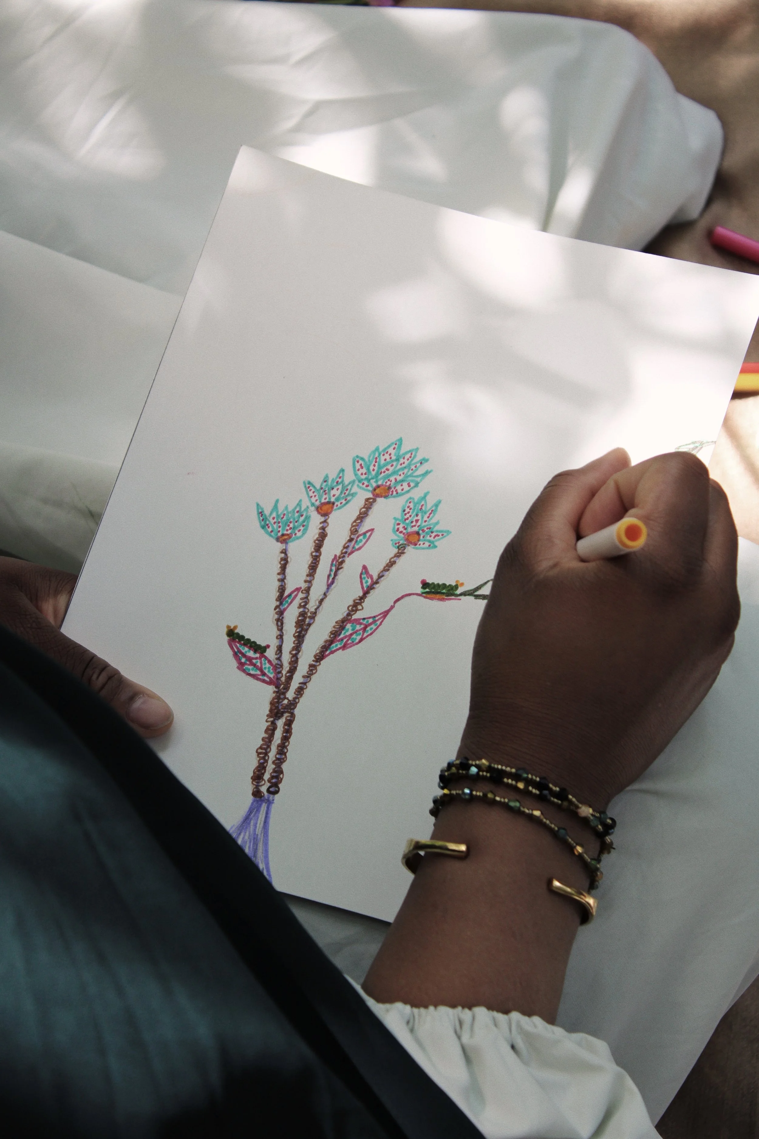 Person drawing colorful flowers and leaves on white paper with a yellow and white marker, wearing multiple bracelets on their wrist.
