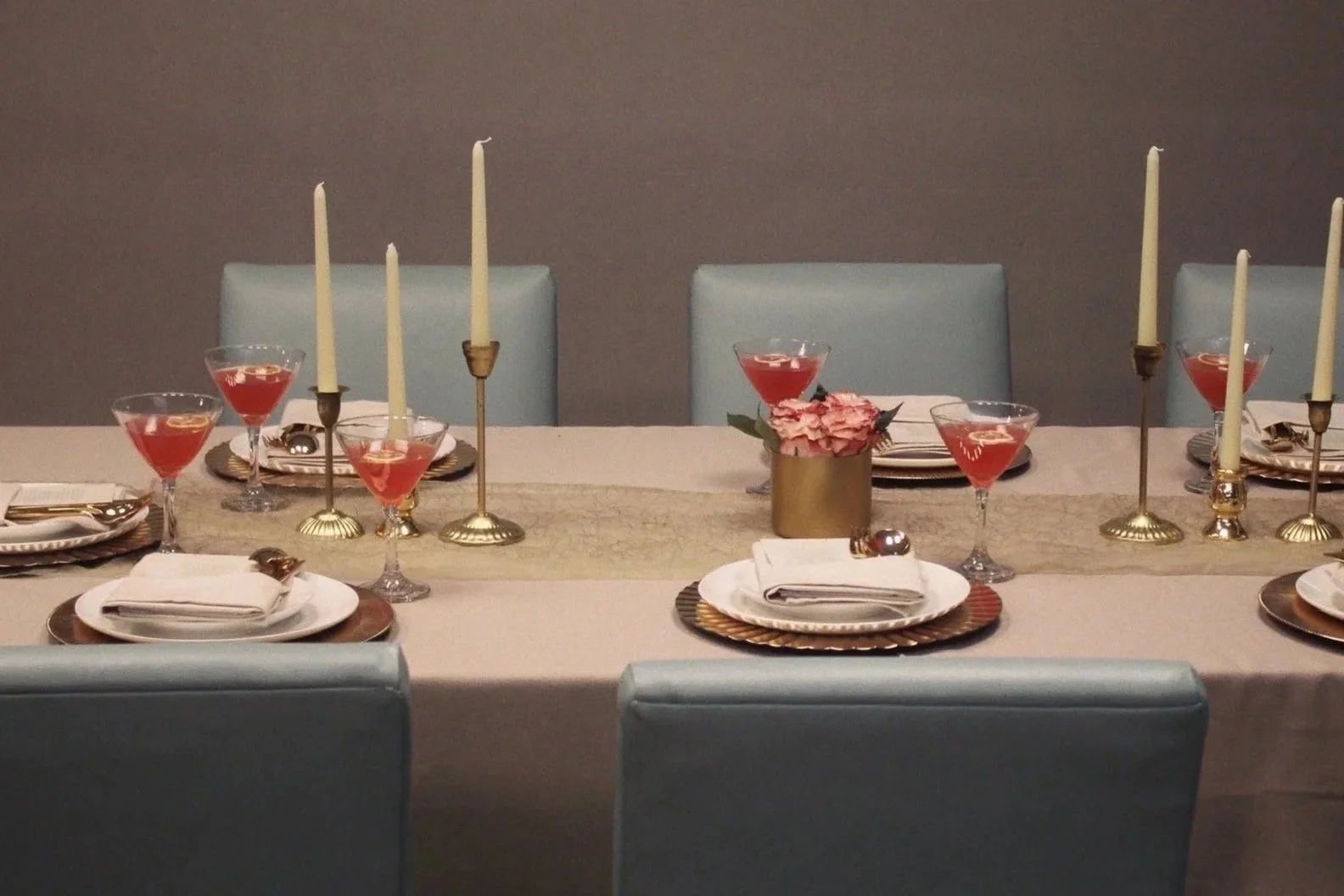 Elegant dining table setup with candles, pink drinks, plates, and silverware, decorated with pink flowers in a gold vase.