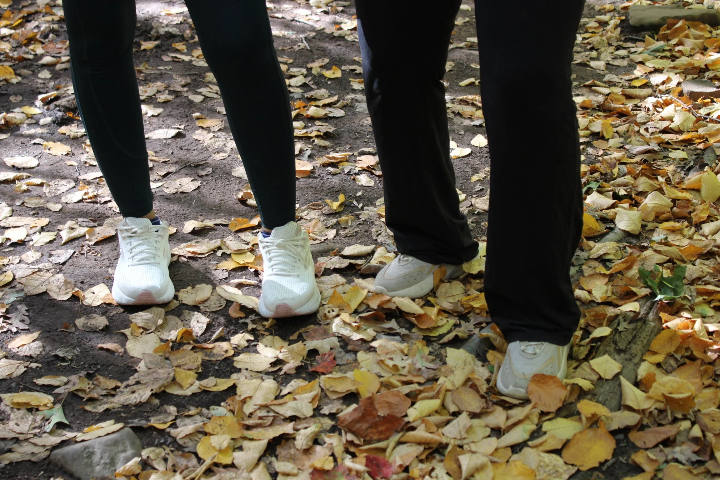 Two people walking on a leaf-covered path in a park or forest, wearing black pants and white sneakers.