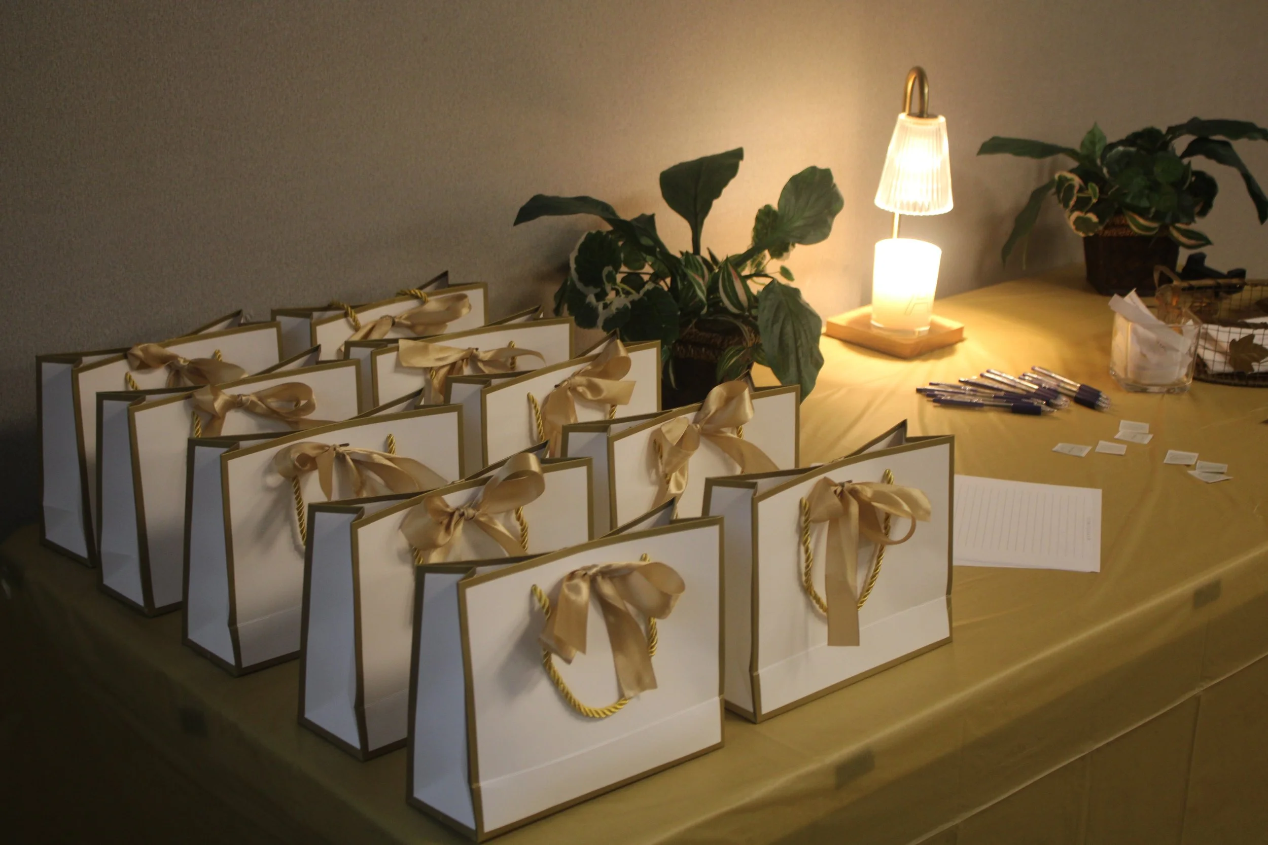 Multiple white gift bags with gold ribbons and handles arranged on a wooden table. The table has plants, a small lamp, pens, small papers, and a notebook.