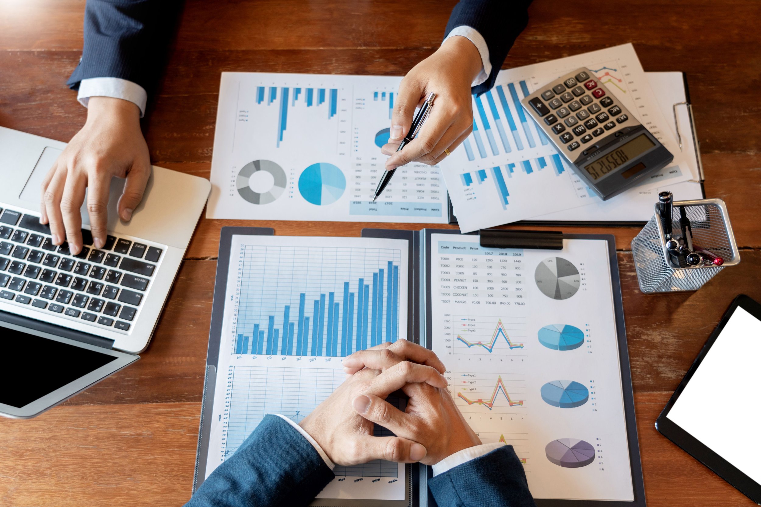 Business professionals reviewing financial charts and graphs on printed papers, laptop, tablet, calculator, and pens on a wooden desk.