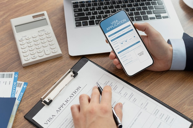 Person filling out a visa application form on a clipboard. Nearby is a laptop, a calculator, airline tickets, and a smartphone displaying a visa application page.