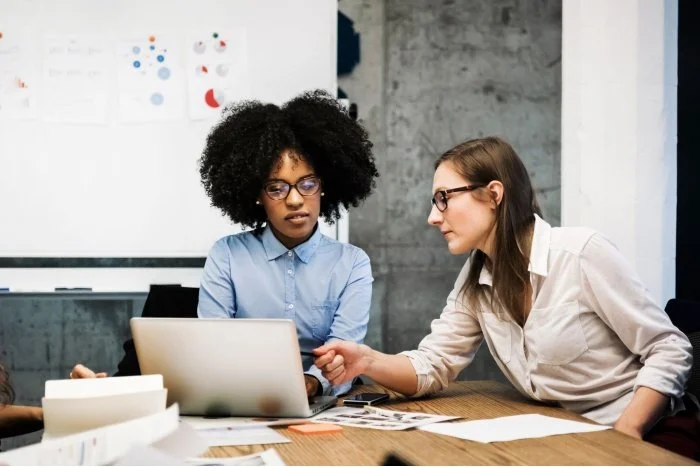 Two women working together at a desk with a laptop, papers, and a whiteboard in the background.