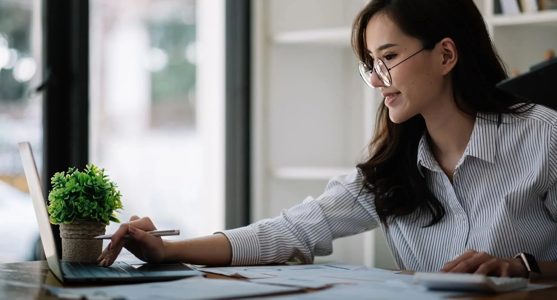 Woman in glasses working on laptop at a desk with a small potted plant