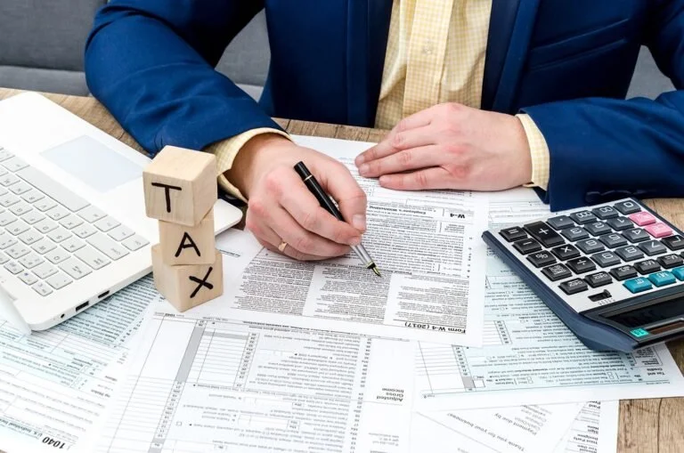 Person in business attire reviewing documents at a desk with a calculator, a laptop, and wooden blocks spelling 'TAX'.