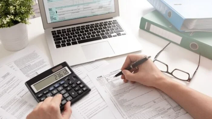 Person working with financial documents, calculator, laptop, glasses, and binders on a white desk.