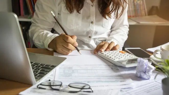 Woman working at desk with laptop, calculator, papers, glasses, and coffee mug.