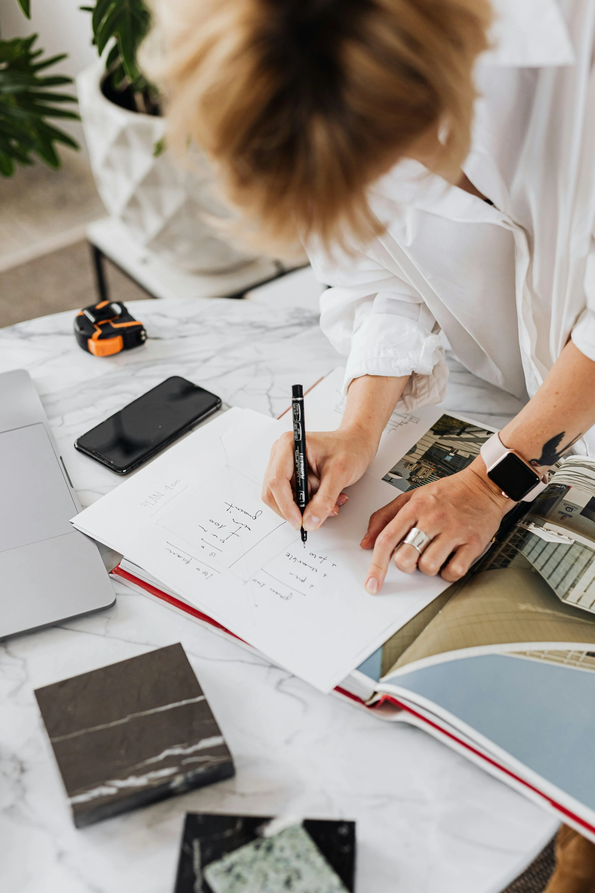 A person with light brown hair working at a white marble table, writing on a sketchbook with a black marker. The table has various items including a black smartphone, a laptop, a tape measure, color swatches, and architectural photos. A potted plant is visible in the background.