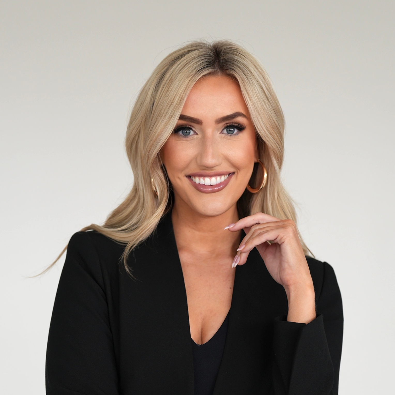 A woman with blonde hair, blue eyes, and hoop earrings, smiling and dressed in a black blazer, posing against a plain light background.