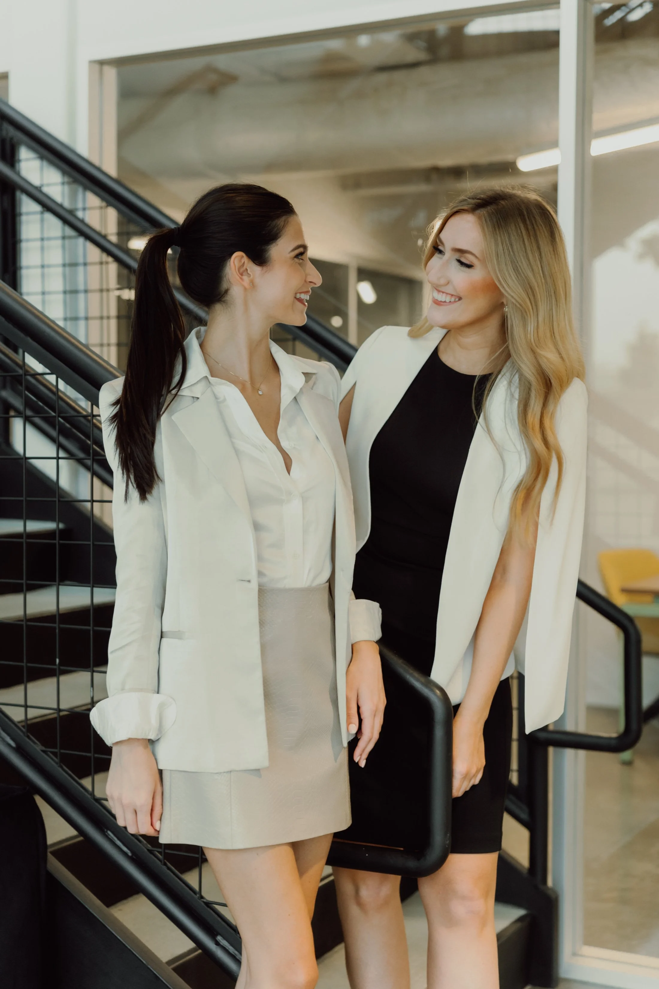 Two women in business attire sharing a moment of happiness on a staircase inside an office building.