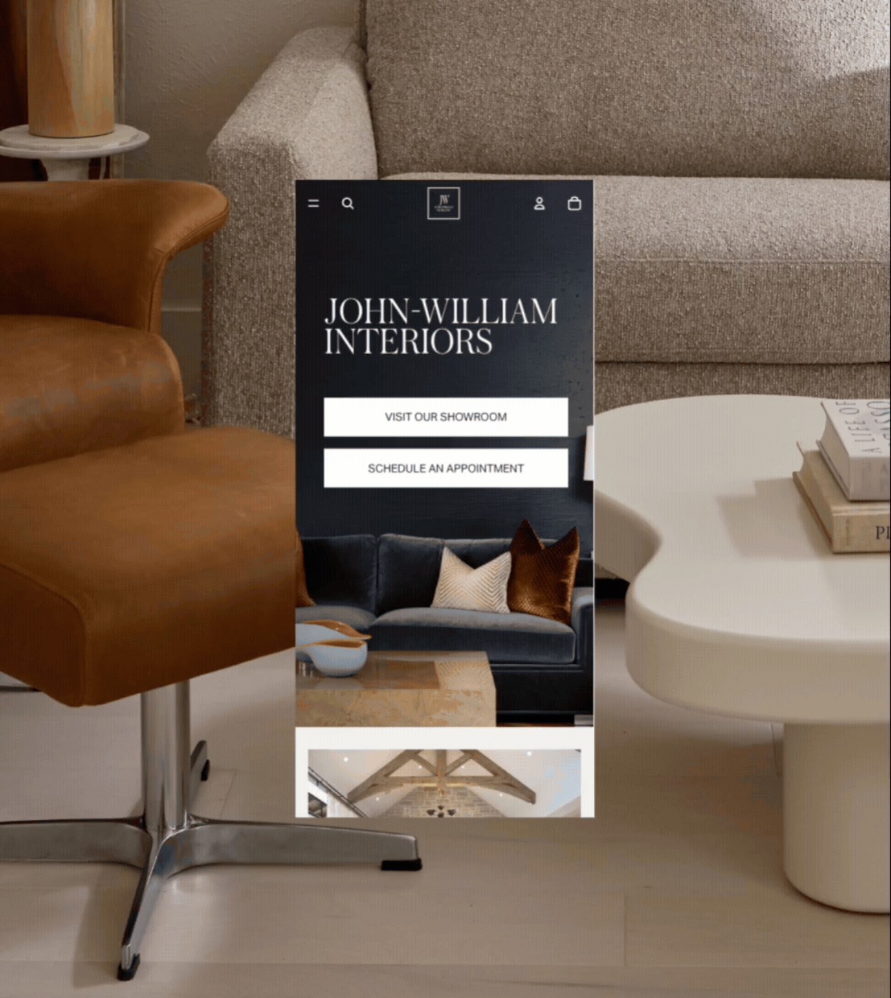 Mobile phone displaying an interior design company's website called John-William Interiors, showing options to visit showroom and schedule an appointment, in a living room with beige sofa, leather chair, round white table with books, and a beige side table with a lamp.