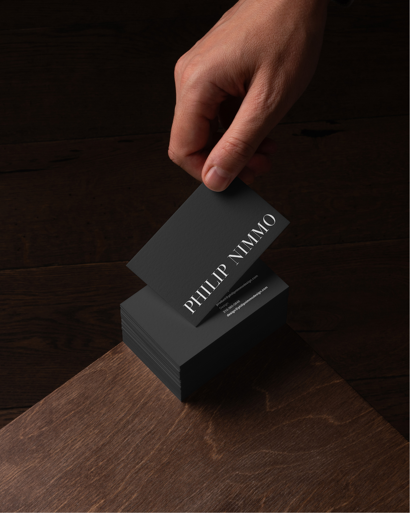 A hand is stacking black business cards with white text that reads 'PHILIP NIMMO' on a dark wooden table.