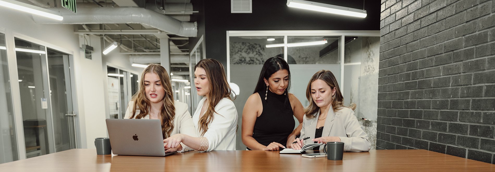 Four women gathered around a table in a modern office space, looking at a laptop and notes while collaborating.