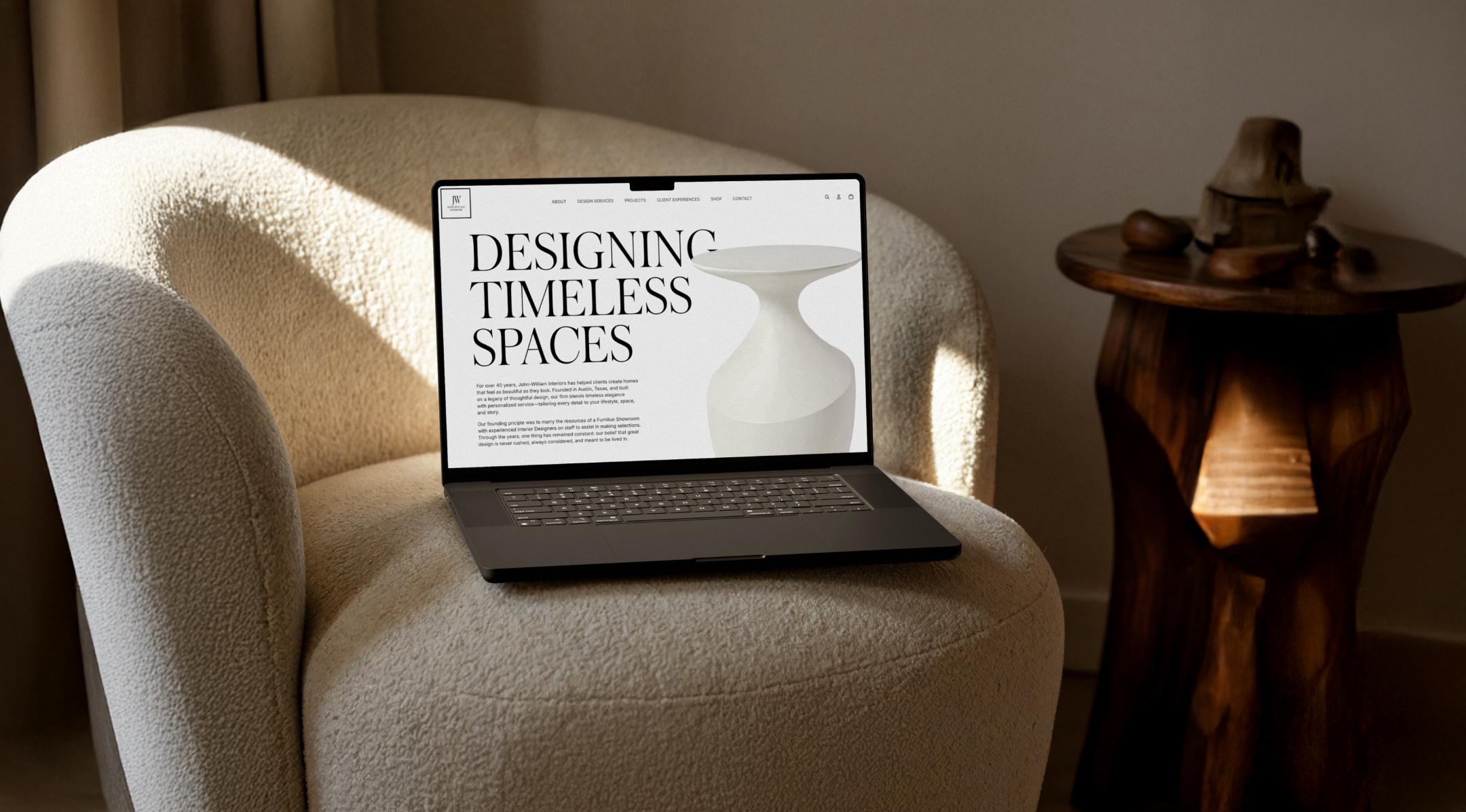 Open laptop displaying a website with the headline 'Designing Timeless Spaces' on a beige armchair, next to a wooden side table with carved wooden objects.