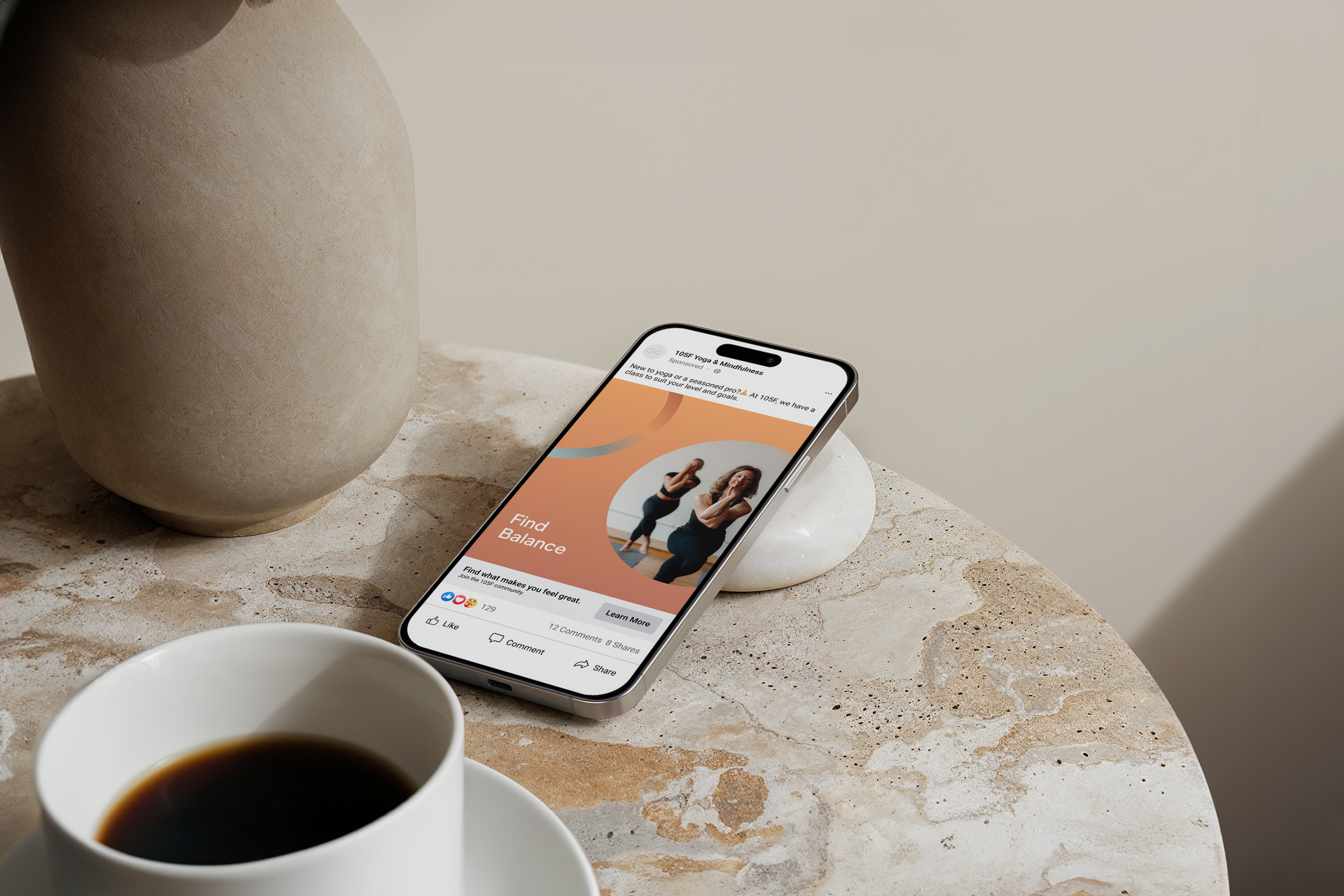 A smartphone on a marble surface displaying a fitness app with a photo of two women doing yoga poses, partially obscured by a white coffee cup, and a large beige vase in the background for 105F hot yoga and pilates in Chicago.