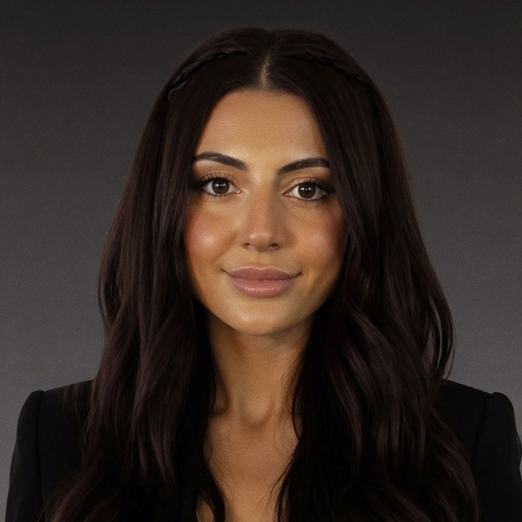 A woman with long dark wavy hair and a natural makeup look, wearing a black blazer, facing the camera with a neutral expression against a dark background.