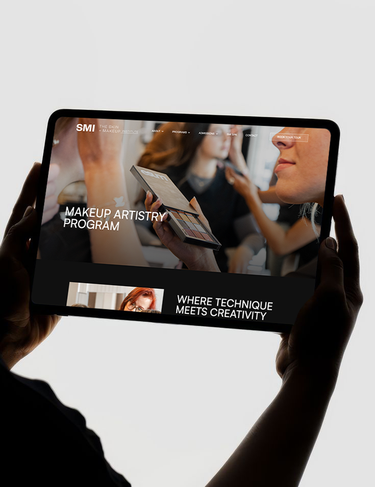 A person holding a tablet displaying a beauty school's makeup artistry program webpage with images of makeup application and students.