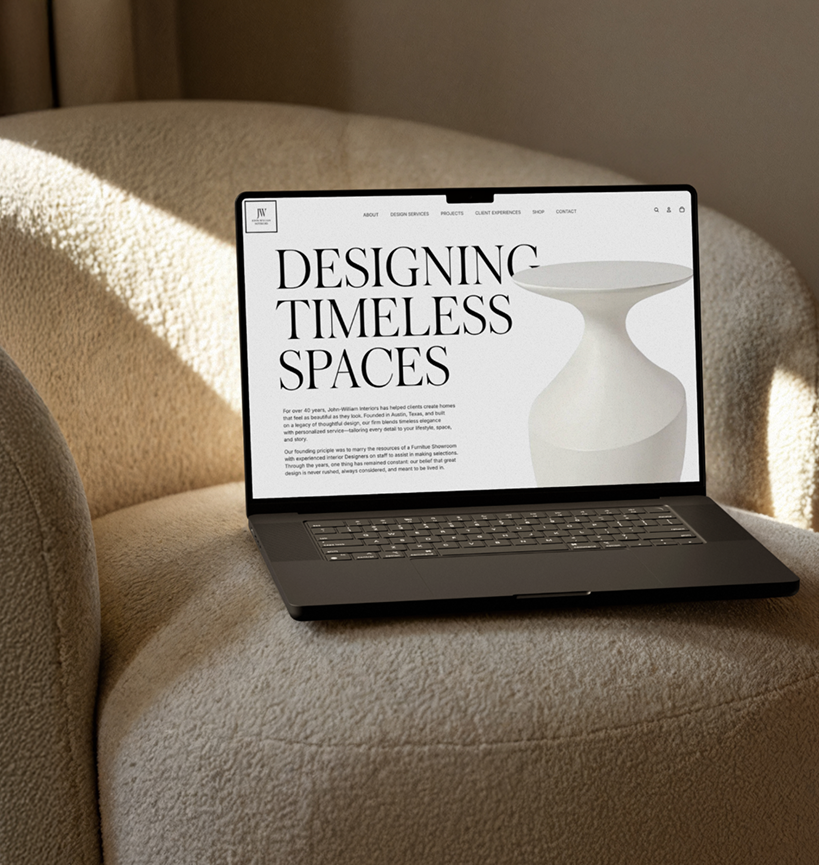 Laptop on a beige armchair displaying a website with the headline "Designing Timeless Spaces" and an image of a white vase for a luxury interior design and fine furnishings showroom in Austin, Texas.