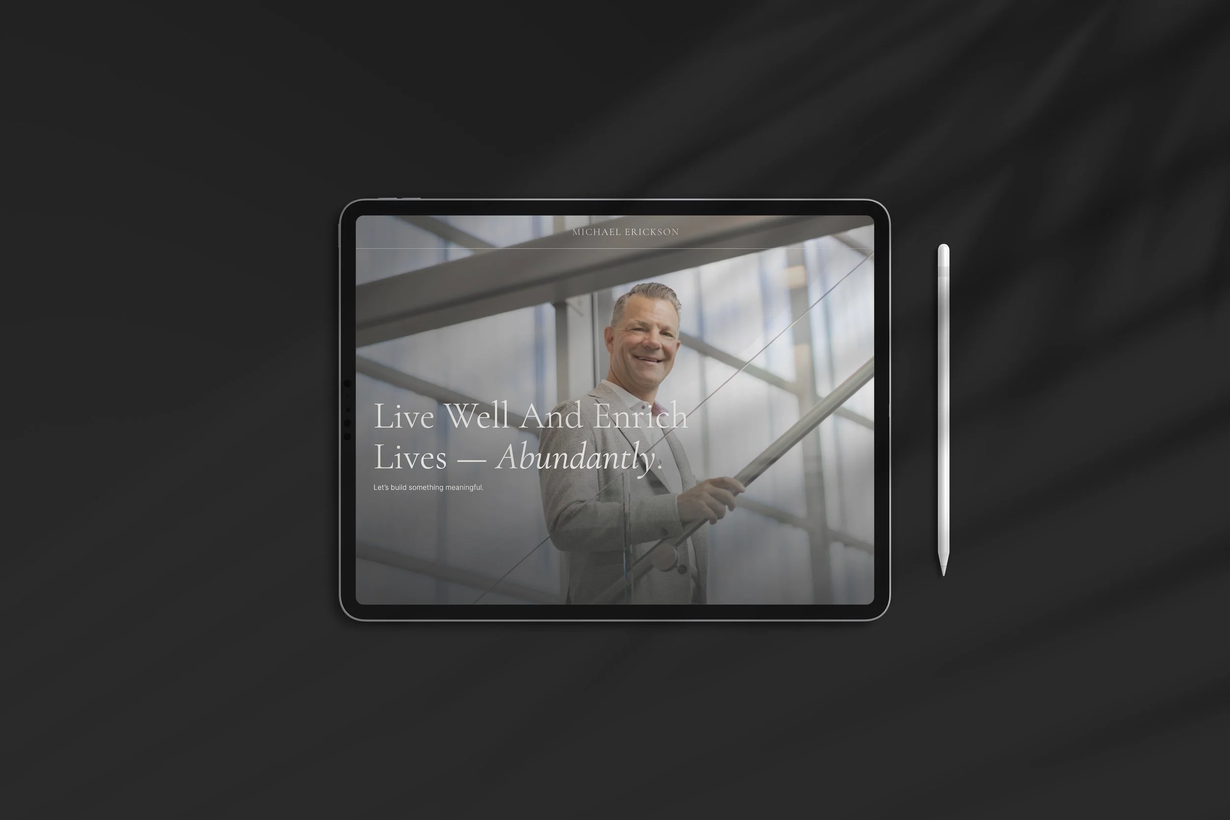 Digital tablet showing a professional man smiling, with overlaid text: "Live Well And Enrich Lives - Abundantly" and a website header "Michael Erickson." The tablet is placed on a dark surface next to a stylus.