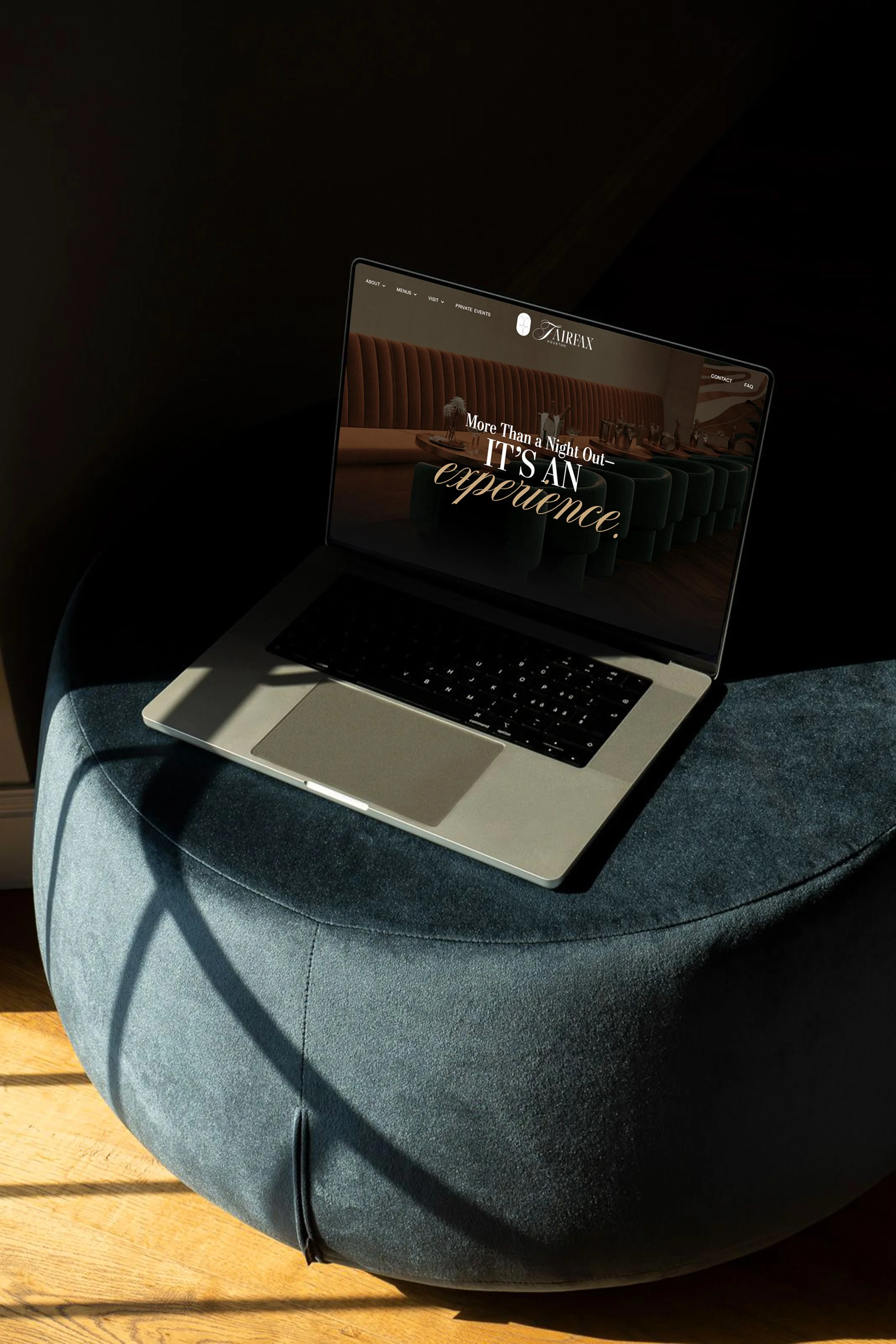 The laptop screen displays a website for a hotel or restaurant with the text, "More Than a Night Out – It's an Experience" and a photograph of a dining area with a long table and dark green chairs for Fairfax Cocktail Bar in Houston, Texas.