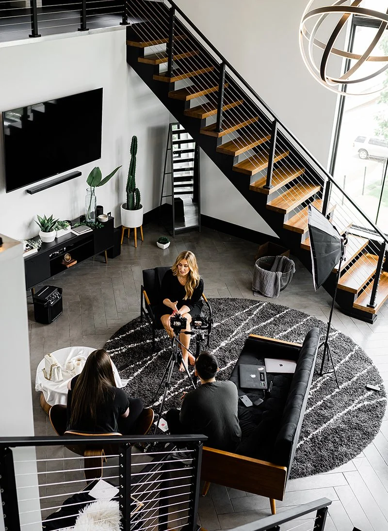Indoor scene of a woman being interviewed on a black sofa on a circular black and white rug, with two people recording with a camera and a laptop, in a modern living room with a staircase and a large window.