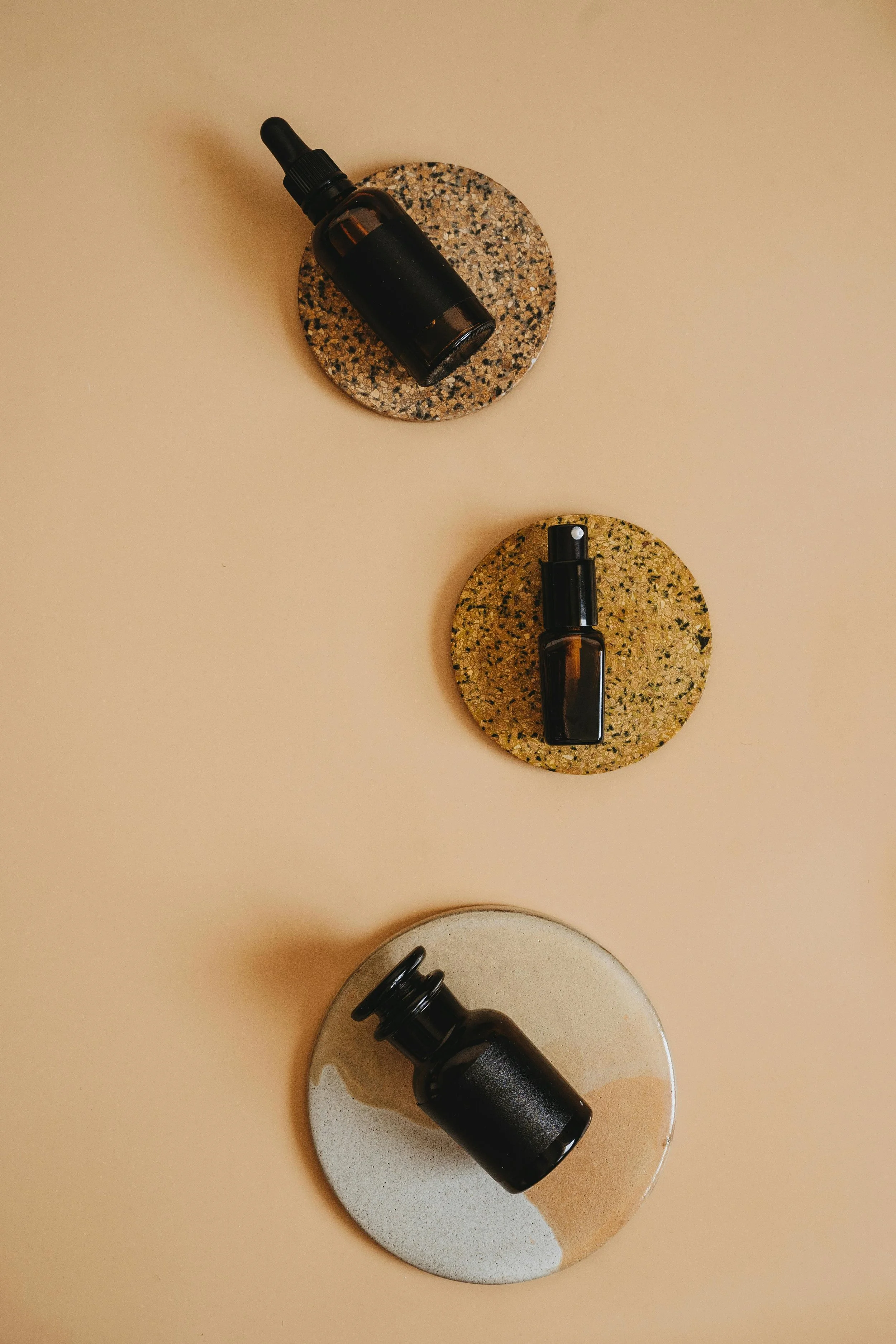 Various small glass bottles with droppers placed on circular stone and ceramic coasters on a beige background.