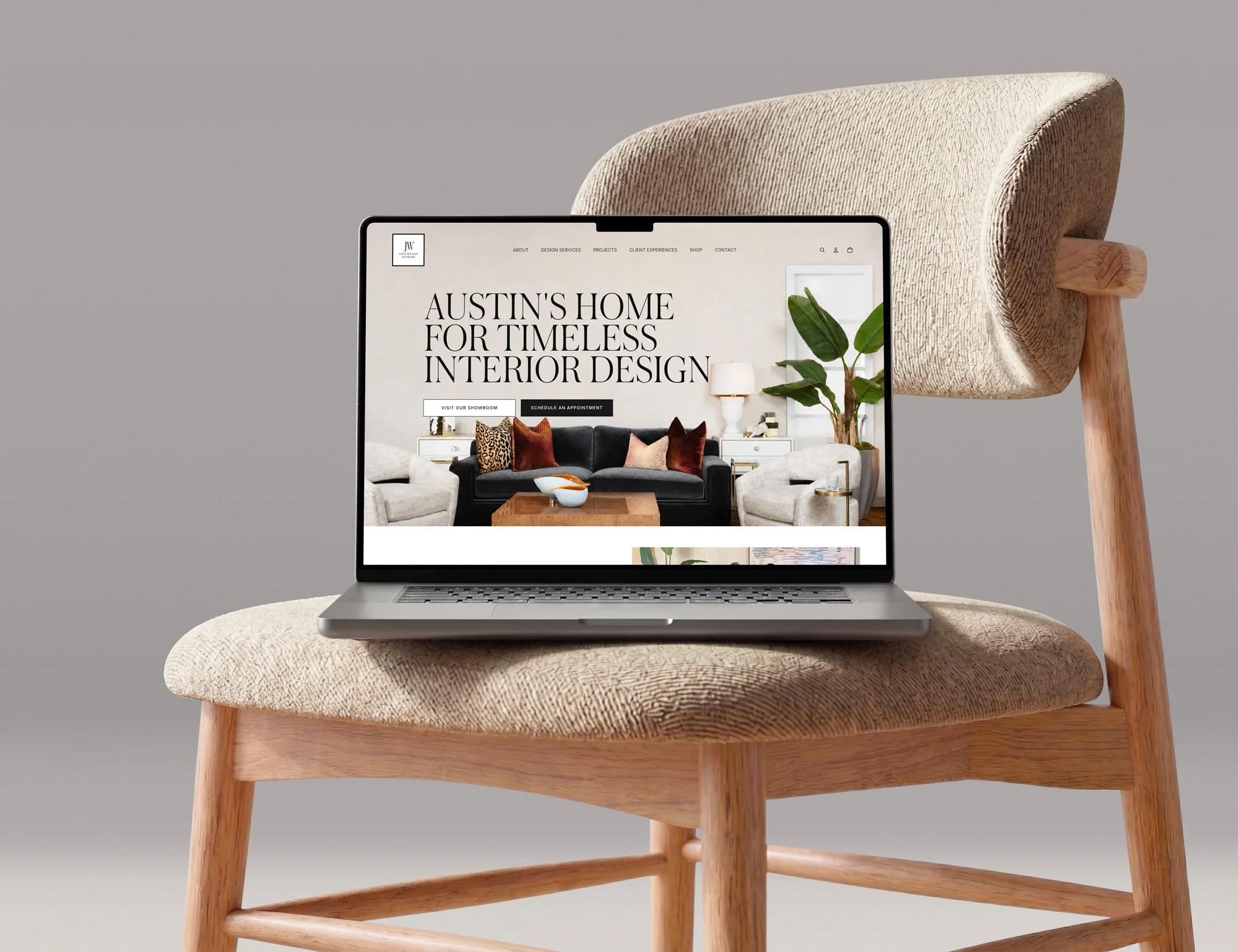A laptop displaying a home interior design website, placed on a beige upholstered chair with wooden legs against a plain gray background for John-William Interiors in Austin, Texas.