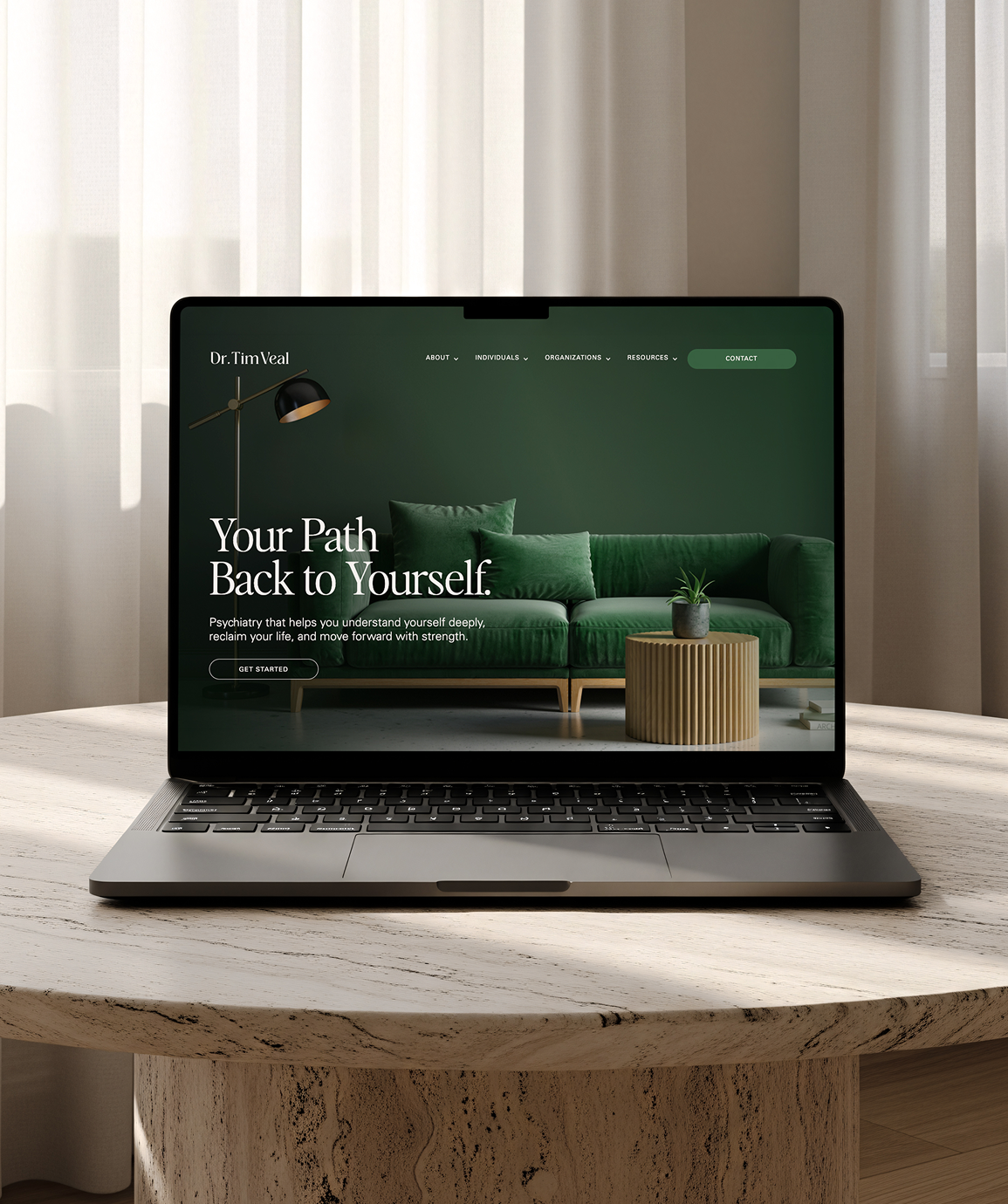 Laptop on a wooden table displaying a mental health website with a green sofa, green pillows, a small plant, and a floor lamp in the background for Dr. Tim Veal in San Diego California.