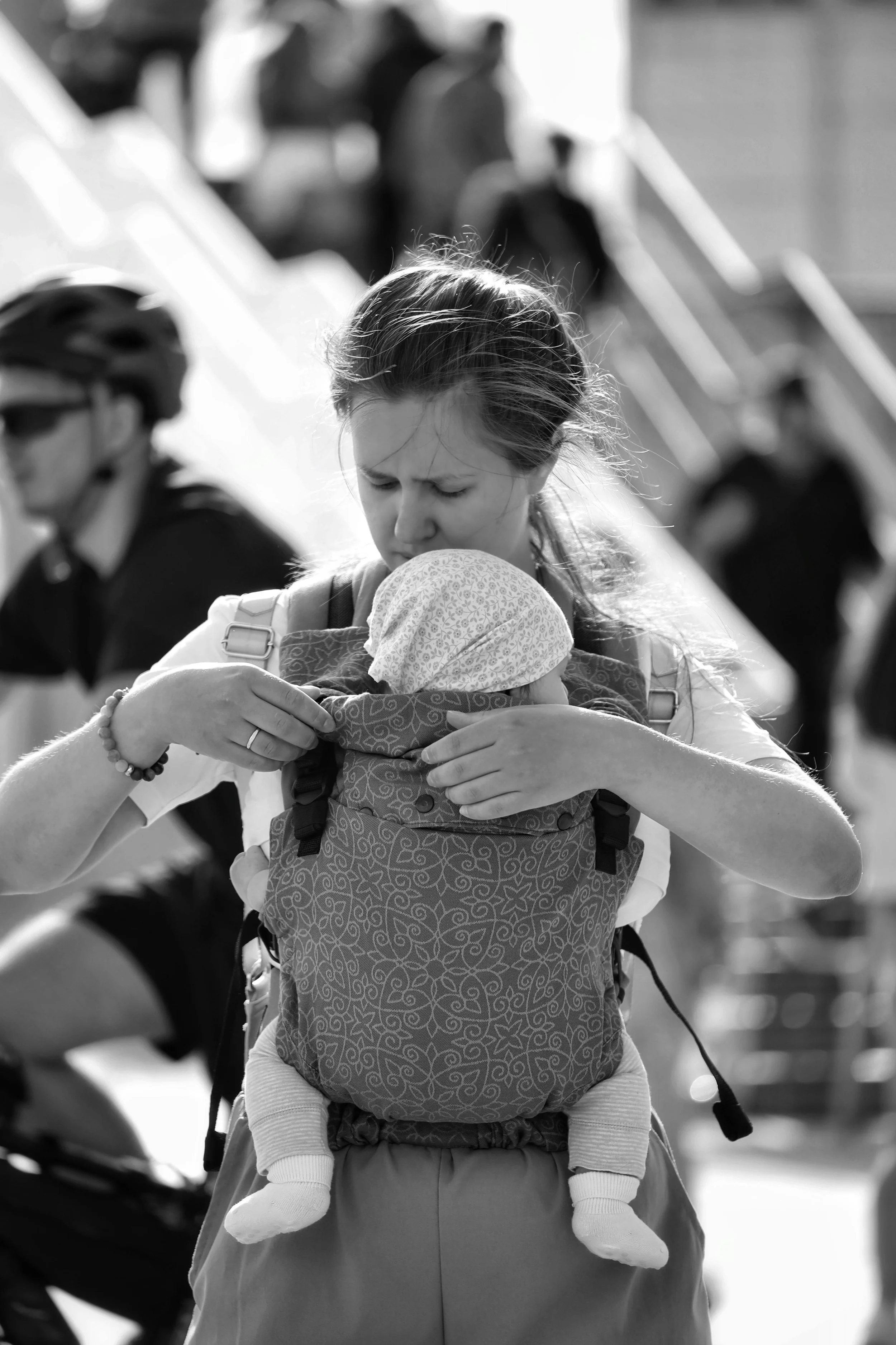 Black and white photo of a woman securing a young child in a baby carrier, with a blurred background of cyclists and an outdoor setting.