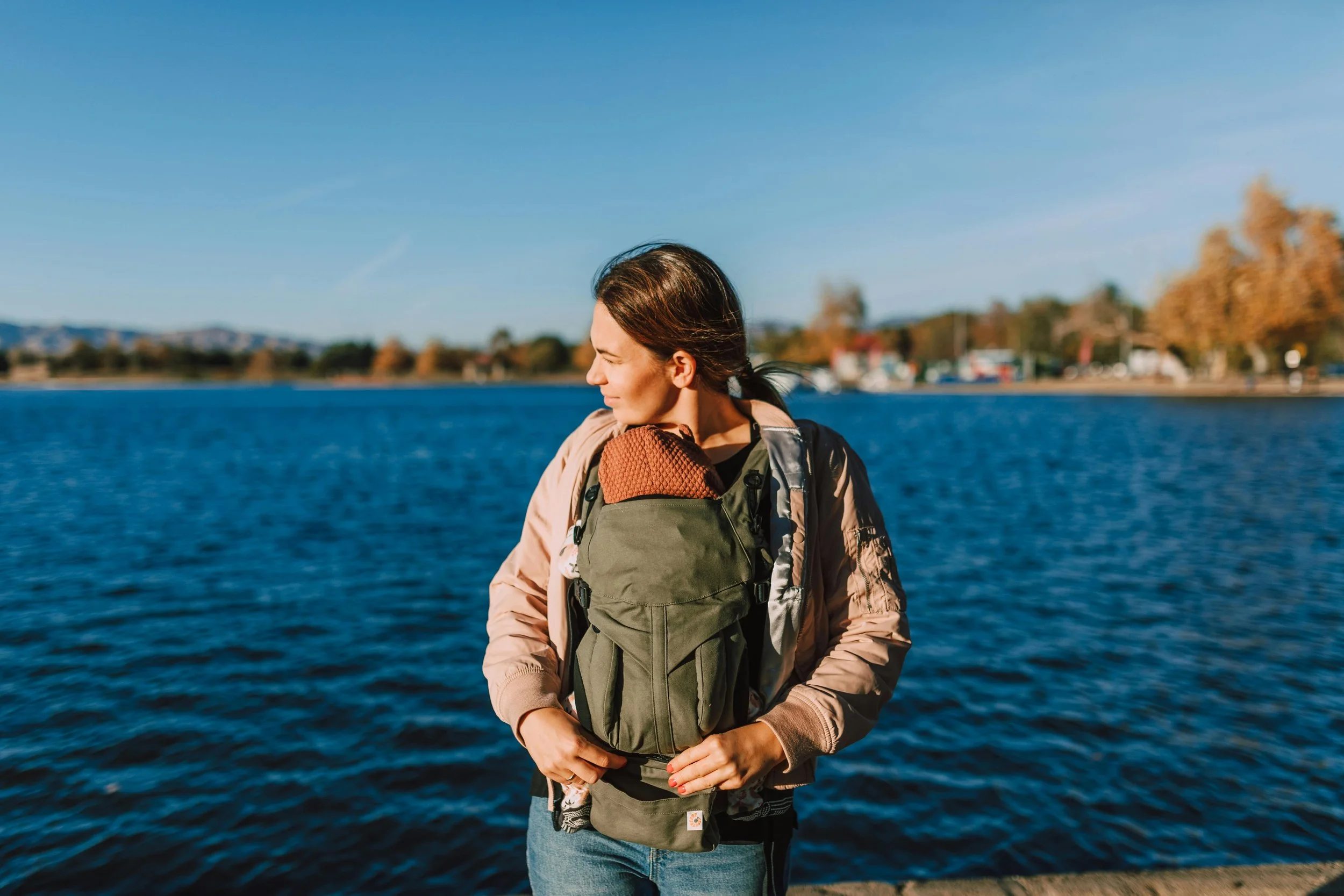 Woman with brown hair, wearing a beige jacket, standing by a body of water with trees and buildings in the background, carrying a child in a green baby carrier with an orange blanket.