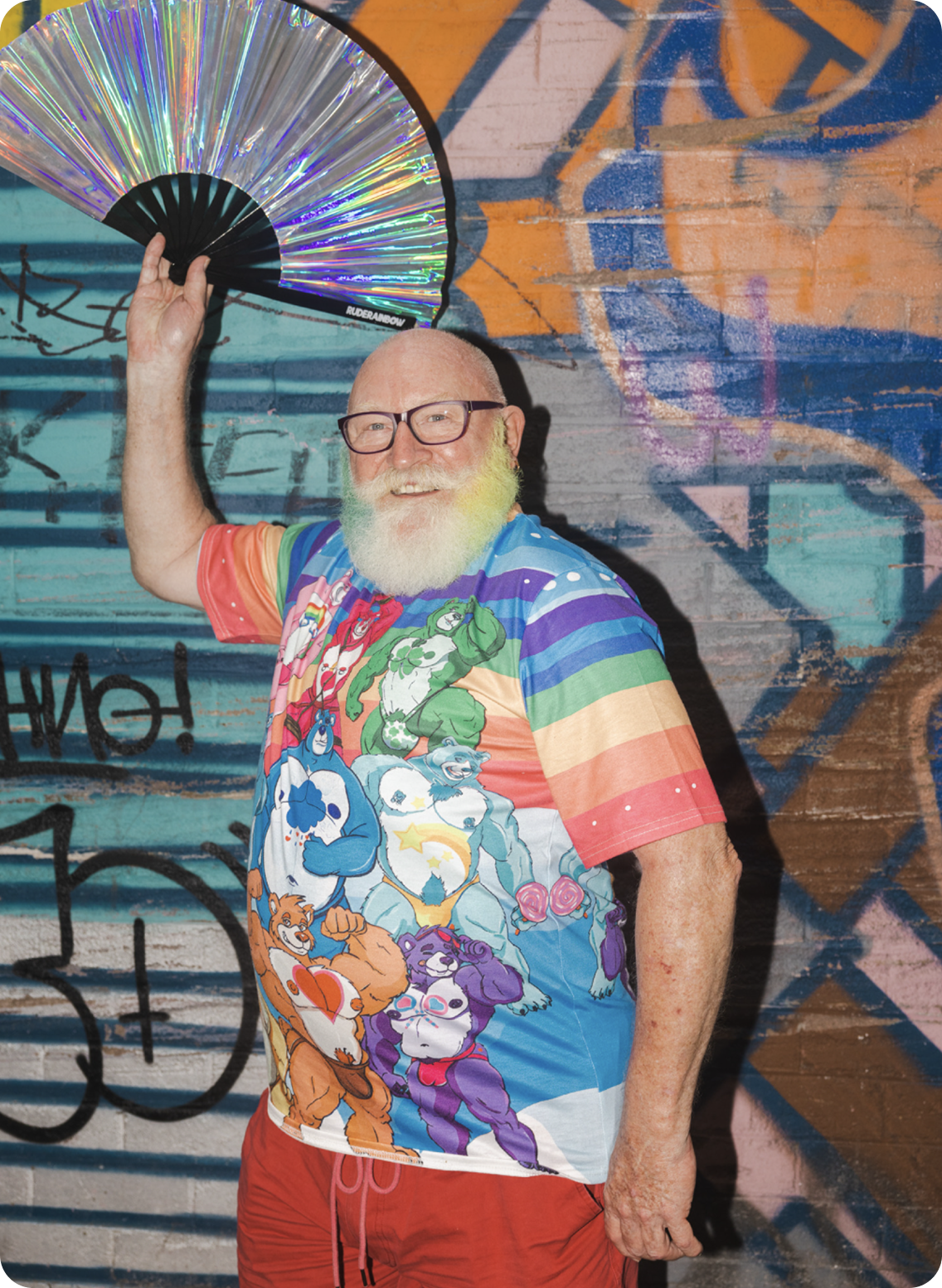 An older man with glasses and a white beard smiling while holding a holographic fan above his head, standing in front of a graffiti-covered wall.