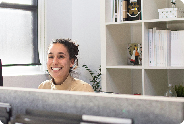 A smiling young woman with dark hair tied back, sitting in an office or study space with white bookshelves in the background.