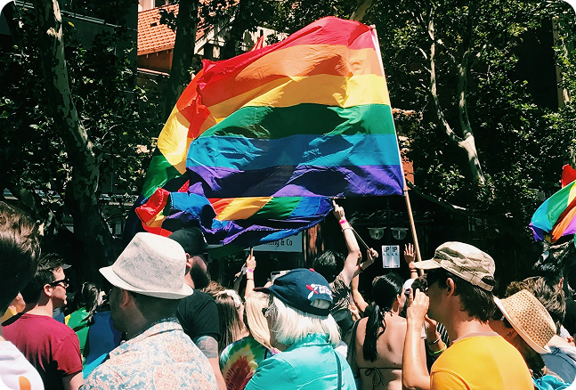 A crowd at a pride parade, with some people holding rainbow flags and wearing colorful clothing and hats.