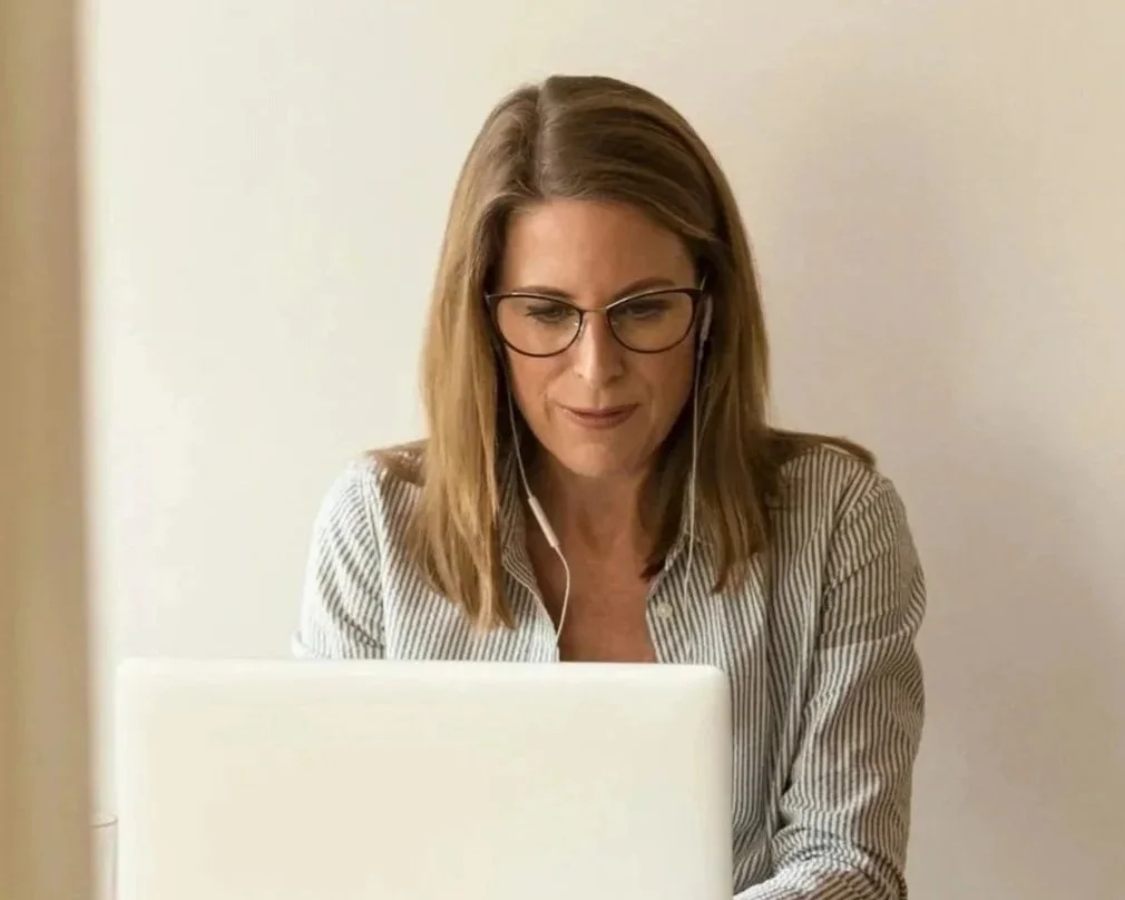 Woman with glasses and brown hair working on a laptop, wearing earphones, in an indoor setting.