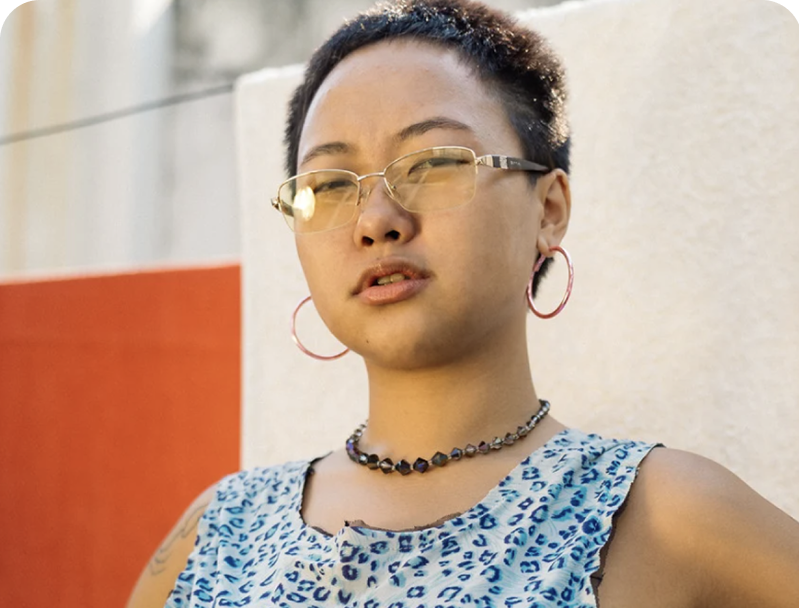 Close-up of a young woman with short black hair, wearing yellow-tinted glasses, pink hoop earrings, a black beaded necklace, and a blue patterned sleeveless top, standing outdoors in front of a beige wall.