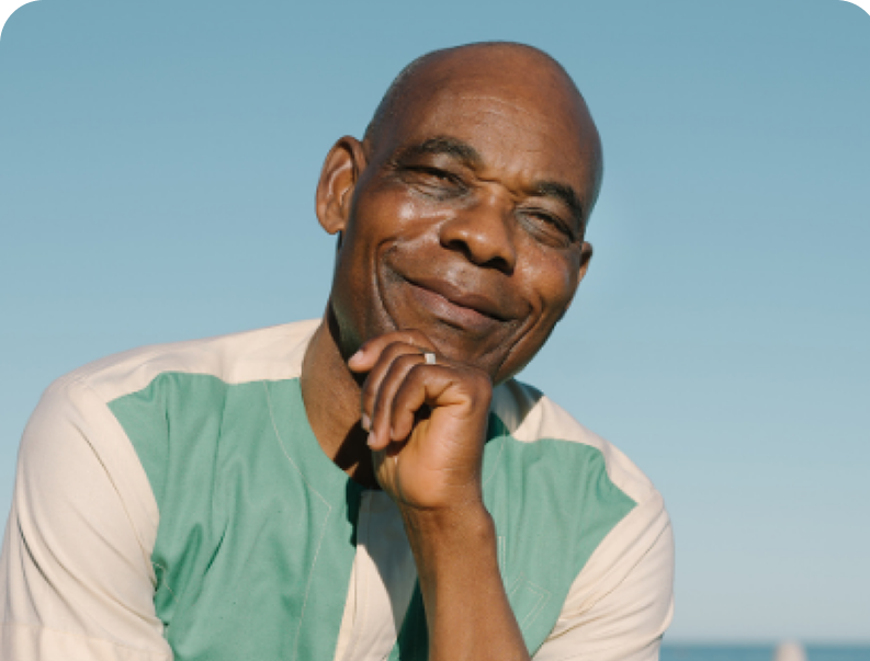 Smiling man with a hand near his chin, wearing a light green and white shirt, against a clear blue sky.