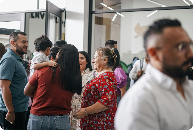 Group of diverse people socializing indoors, with some smiling and chatting, and a woman holding a child on her shoulder.