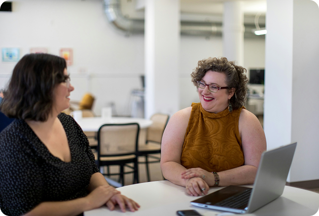 Two women smiling and talking across a table in an office or coworking space, with a laptop open on the table.